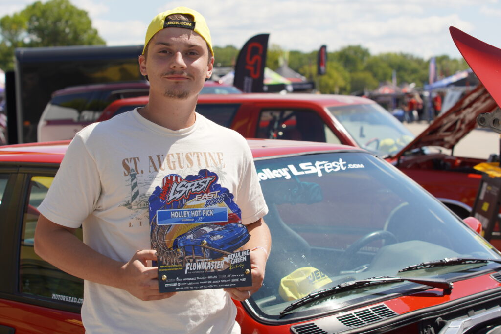 Car enthusiast holding Holley LS Fest East 2025 Holley Hot Pick award in front of red BMW M3 E30 with windshield decal at Holley LS Fest East