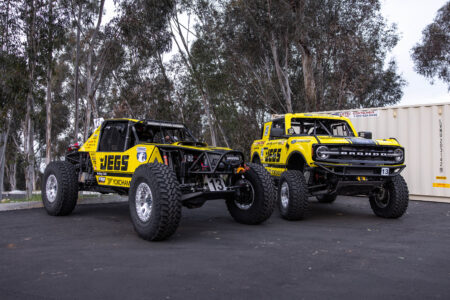 Two bright yellow JEGS-branded off-road race vehicles parked side by side on pavement, including a tube-chassis Ultra4-style buggy and a Ford Bronco race truck, both on large off-road tires with the number 13, surrounded by trees and shipping containers in the background.